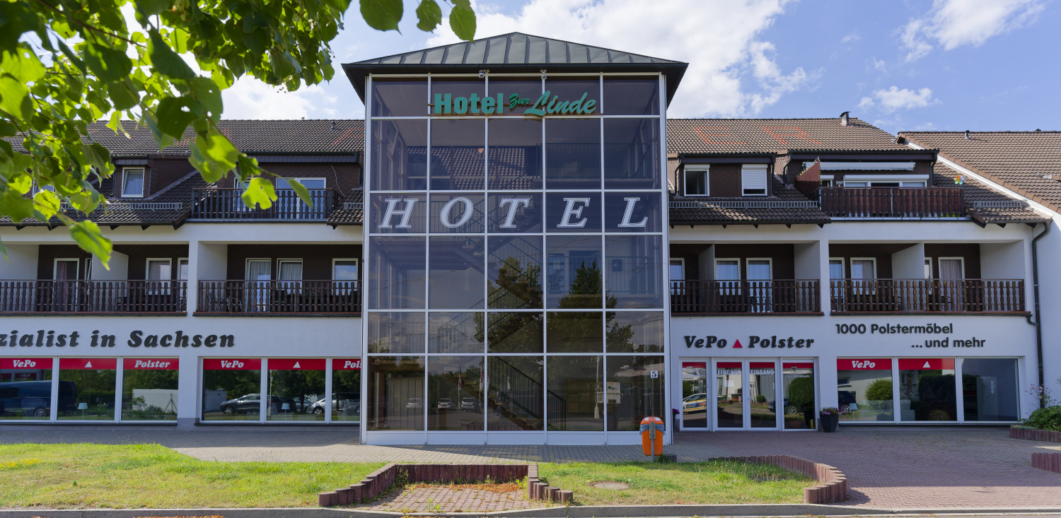 Zeigt das Hotel in der Frontansicht. Es ist ein gläserner Aufgang zu sehen. Das Hotel befindet sich im ersten Obergeschoss. Darunter ist ein Polstermöbelgeschäft eingerichtet und darüber sind Privatwohnungen.