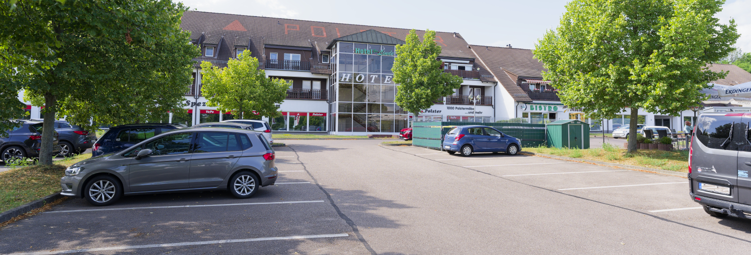 Zeigt das Hotel im Hintergrund. Im Vordergrund sind Parkplätze und einige Autos zu sehen. Es gibt freie Parkplätze.